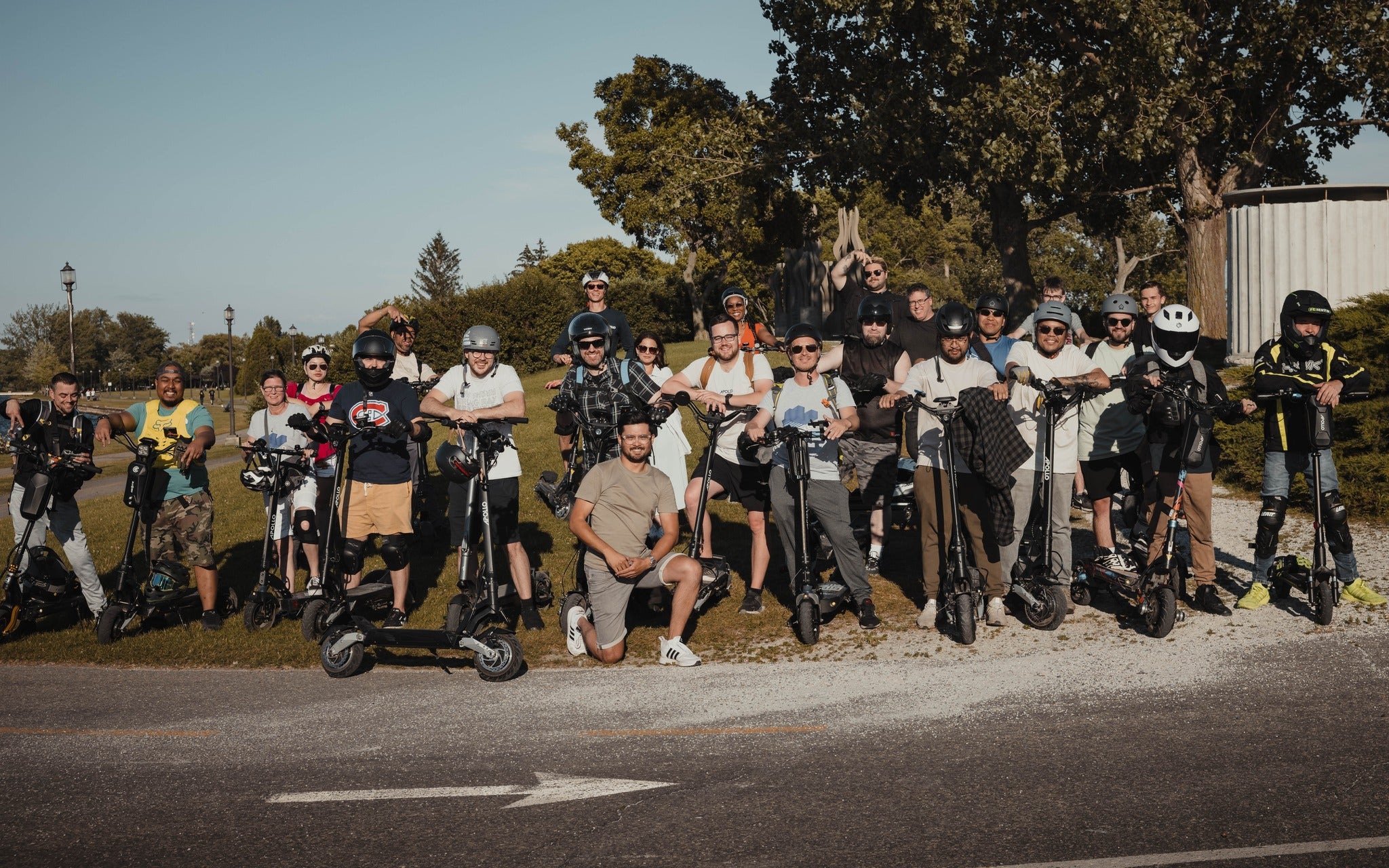 A large group of individuals stand under trees next to electric scooters, ready to ride