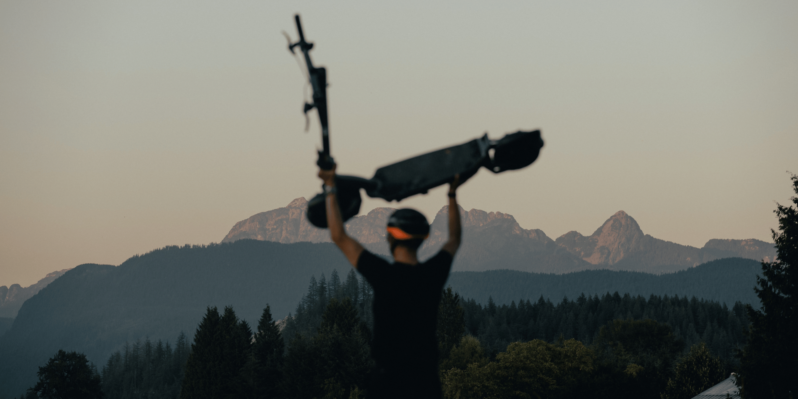 A person holding an electric scooter above their head while viewing mountain peaks in the distance.
