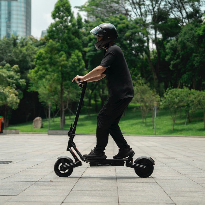 Person riding a black electric scooter in a park with greenery and buildings in the background.