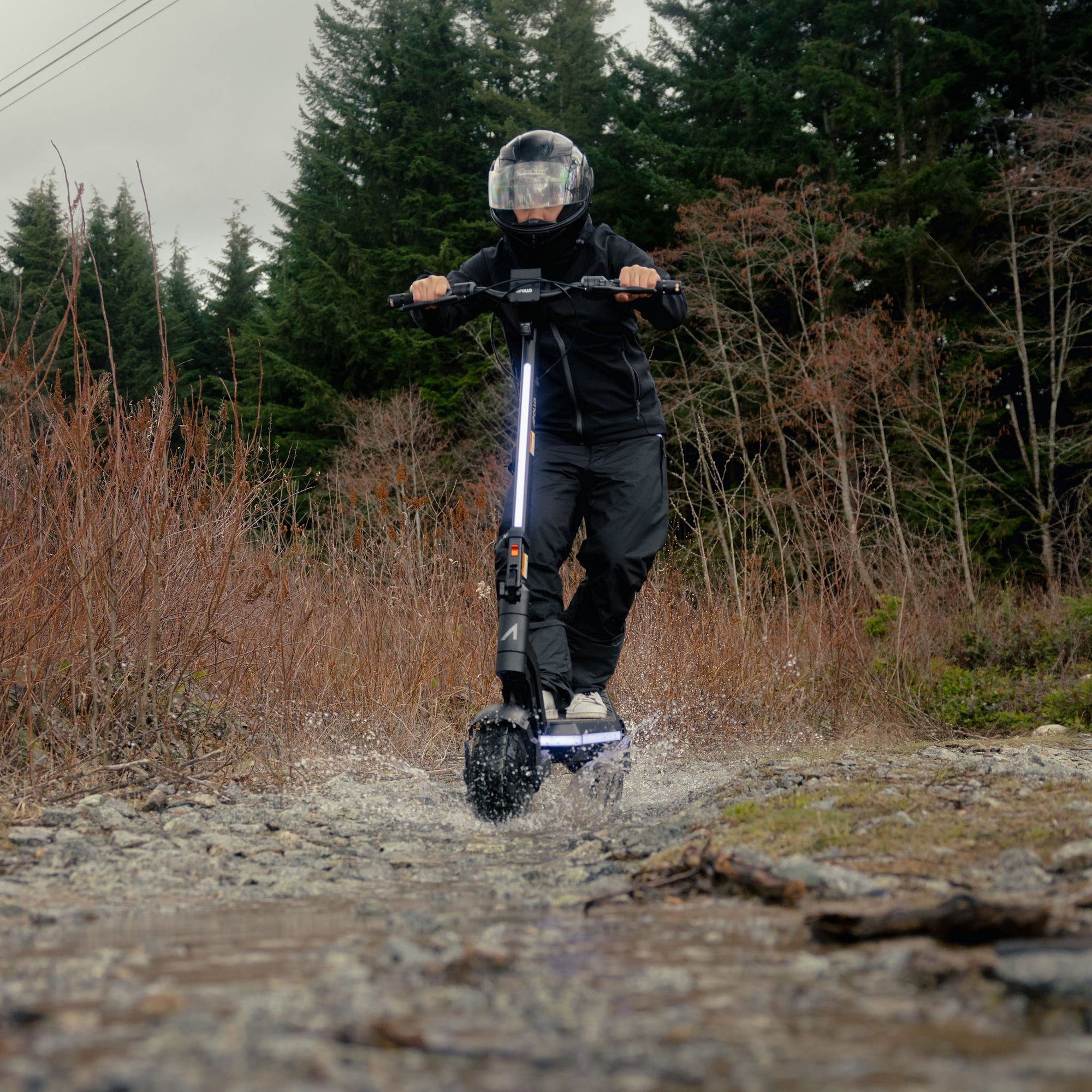 A person riding an electric scooter on a rocky dirt path outdoors, with dry brush and evergreen trees in the background.