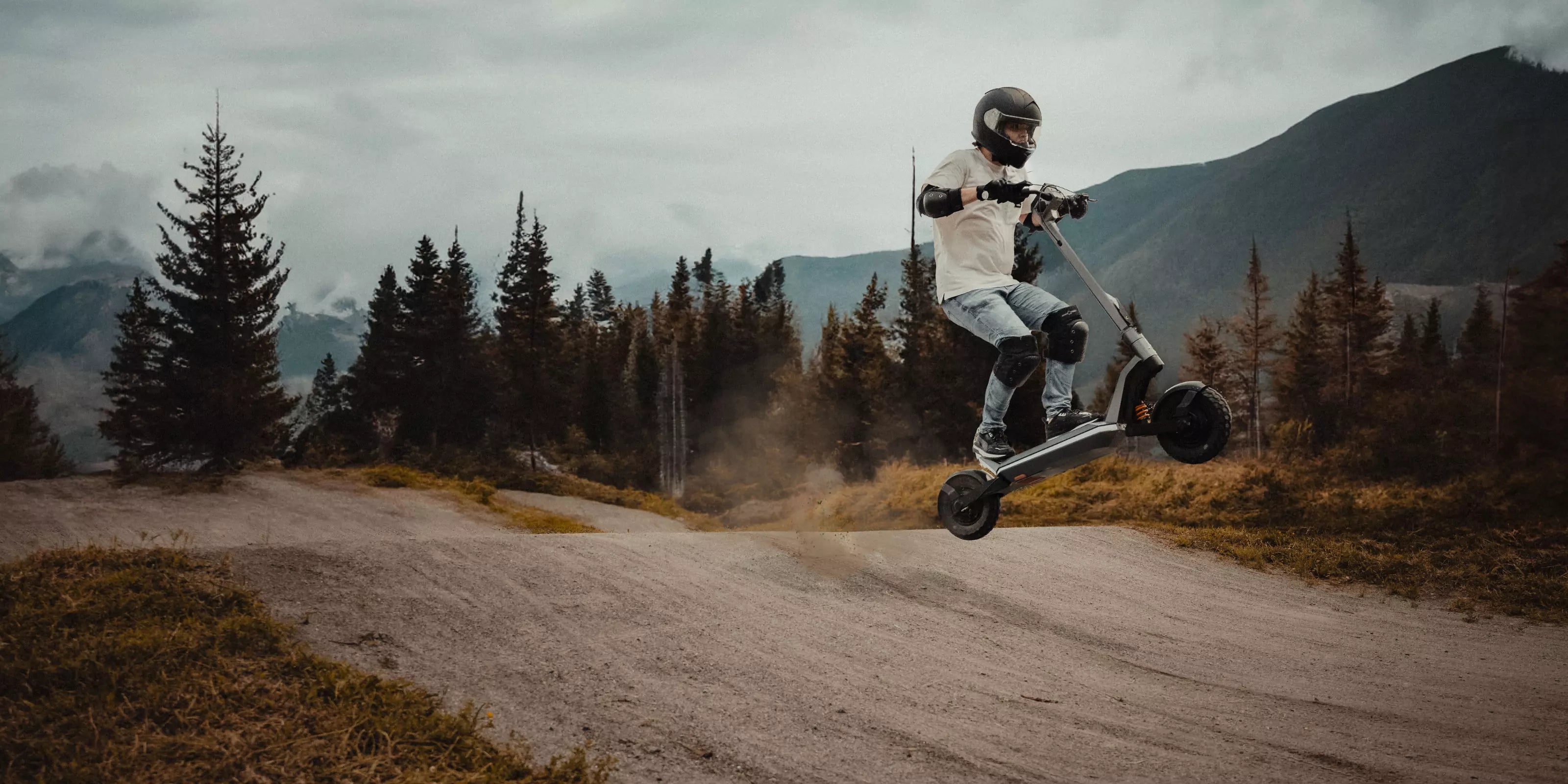 Man riding an electric scooter while jumping off dirt ground outdoors with mountains in the background.