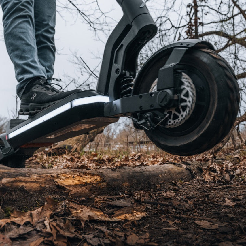 A close-up outdoor shot of a person's foot in a black shoe stepping onto the deck of a black electric scooter, with the rear wheel and fork visible above a forest floor covered in dry leaves and branches.