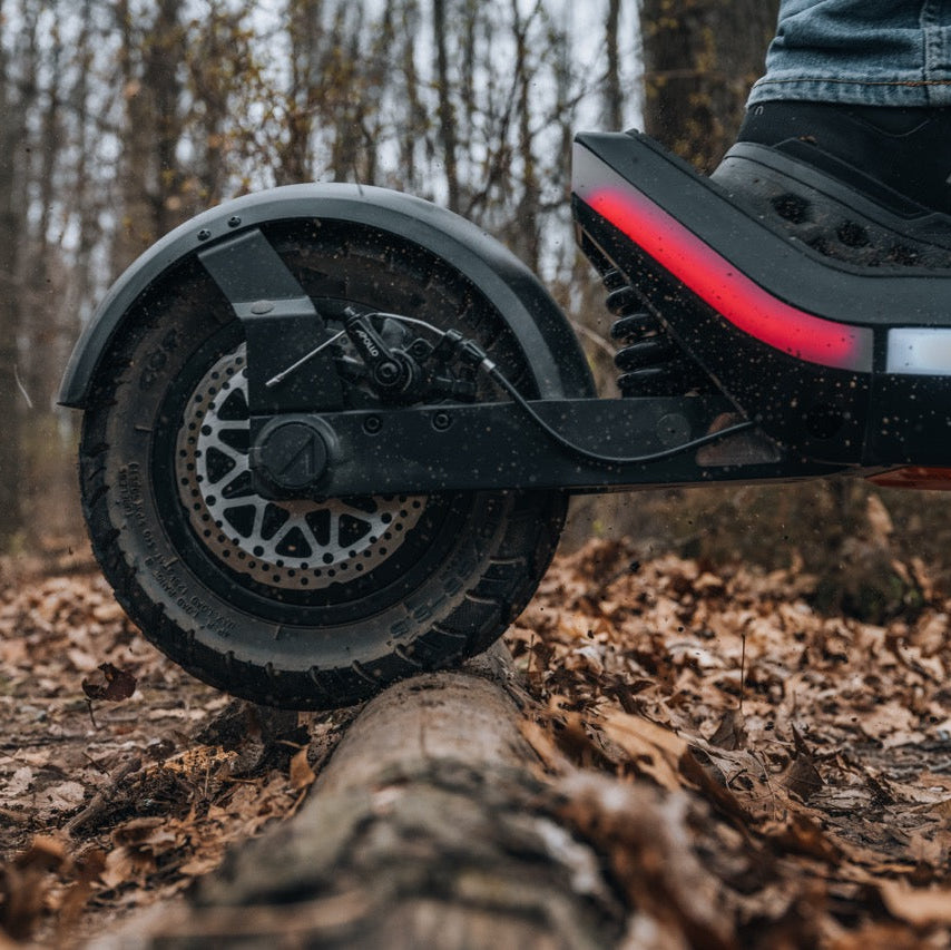 Close-up of a black electric scooter wheel and fender on a leaf-covered log in a wooded outdoor setting, with part of a person's leg and shoe visible on the deck.