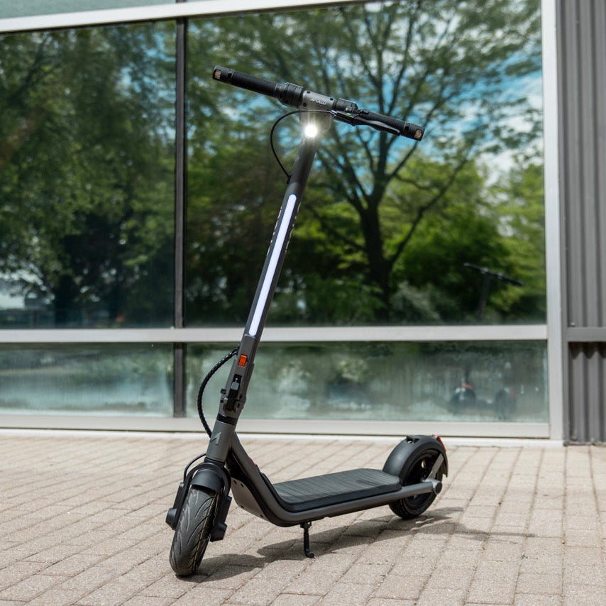 A black electric scooter with a silver frame accent, standing on its kickstand on a paved outdoor area in front of a modern building with large windows.