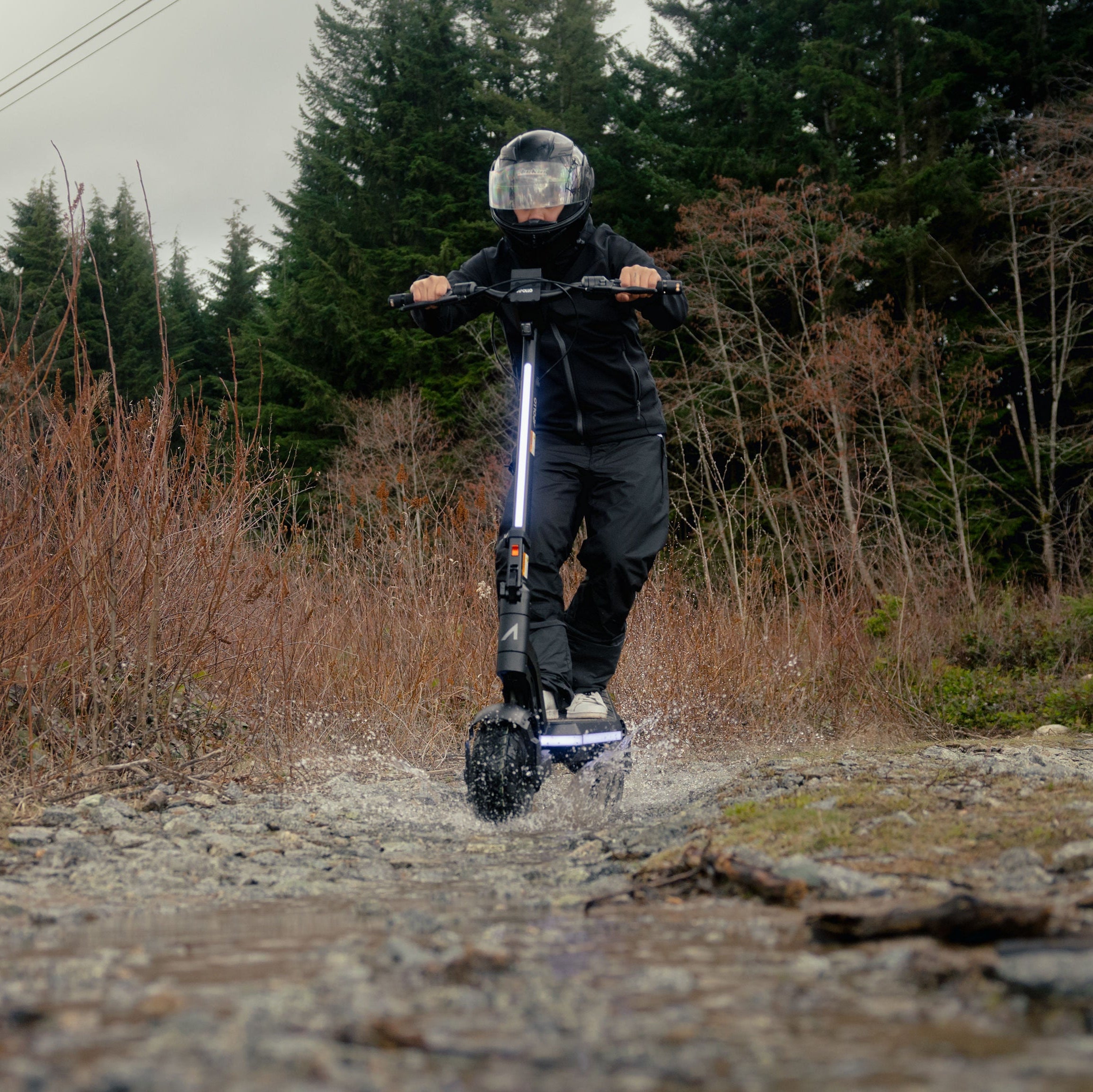 A person riding an electric scooter on a rocky dirt path outdoors, with dry brush and evergreen trees in the background.