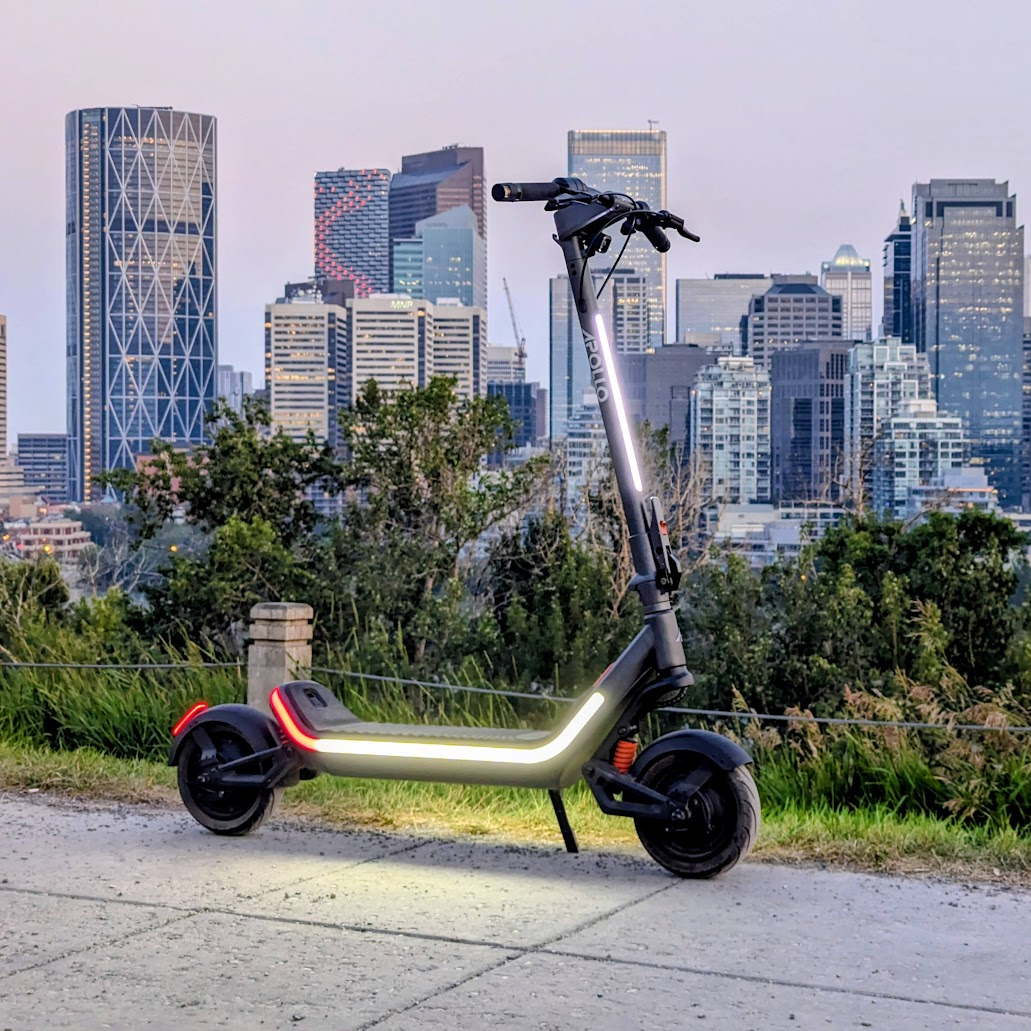 A folding electric scooter with a metallic silver frame and black wheels, parked on a concrete path with a city skyline and green trees in the background.